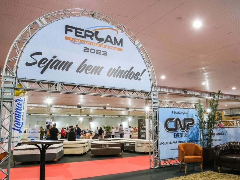 Arco de entrada da Feira Capixaba de Móveis (FERCAM 2023) com a inscrição "Sejam bem vindos!" e estandes de colchões visíveis ao fundo.